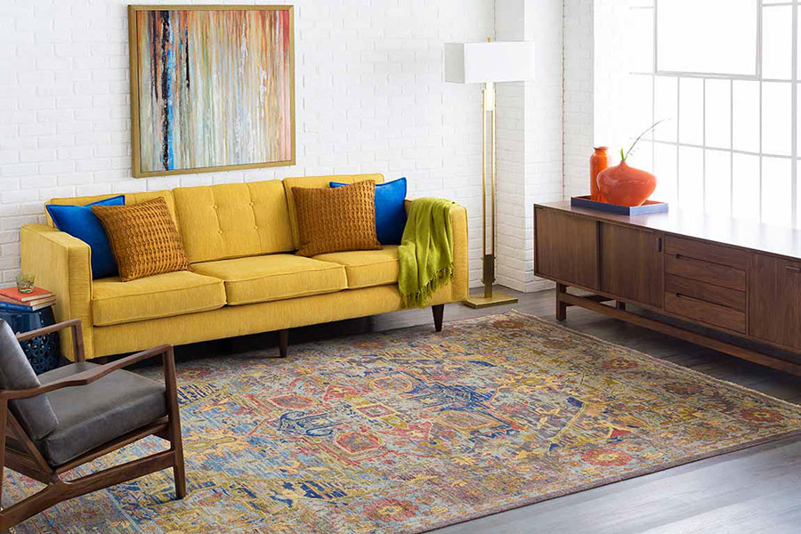 Living room with a yellow cloth couch, blue and gold throw pillows, grey chair on a large area rug and engineered floors.
